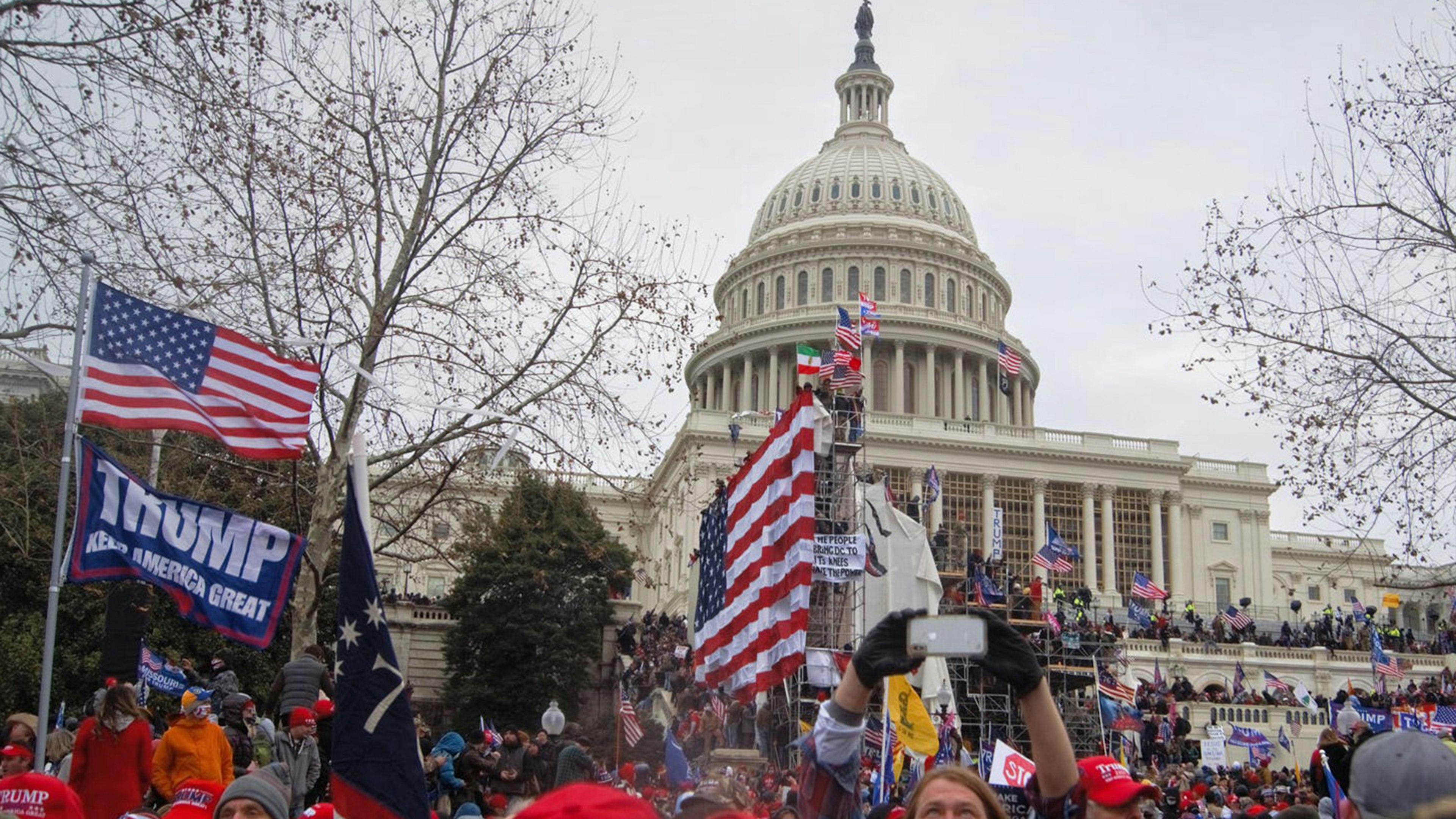 Photo of protesters at U.S. Capitol in Washington, D.C. on January 6, 2021 during insurrection. People stand on stairs outside Capitol Building with American flags and a 'Trump Make America Great Again' flag.