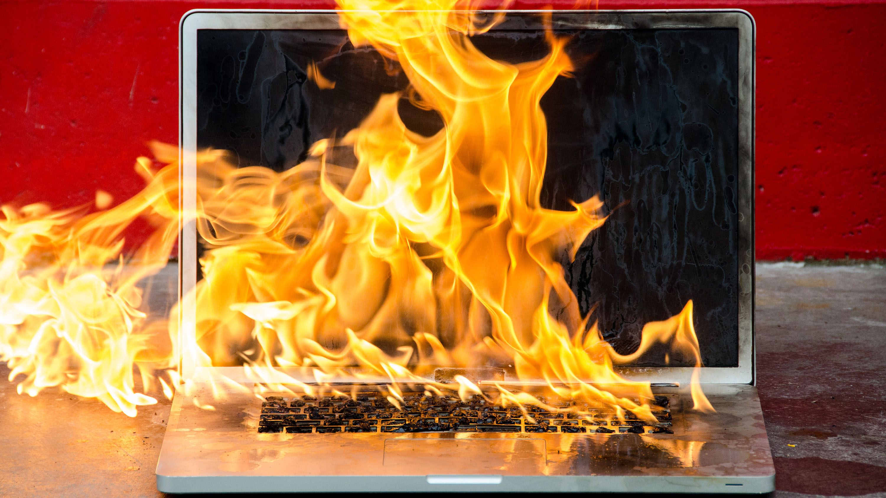 A laptop engulfed in bright orange flames against a red background.