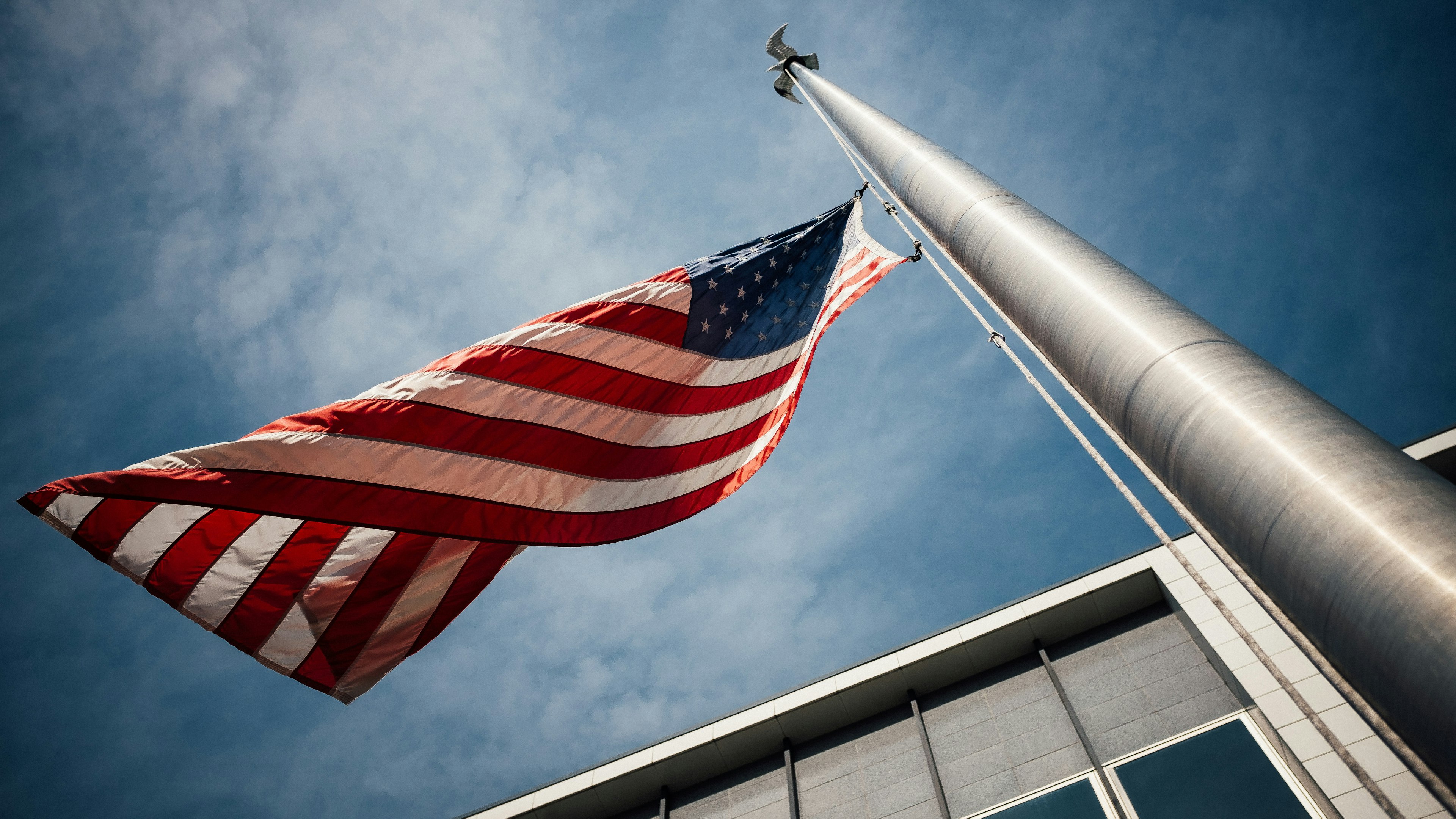 Photo of an American flag waving on a pole with a blue, cloudy sky in the background. Picture is taking looking upward at the flag.